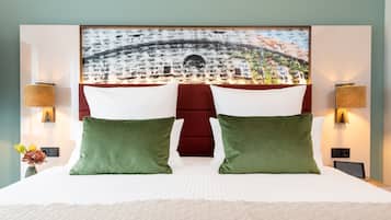 A hotel room with a large bed, two green pillows, a red headboard, and a wall-mounted painting of a cityscape.