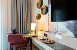 Una habitación de hotel moderna con una mesa redonda, una silla roja, una computadora portátil, una lámpara, un televisor y un plato decorativo con comida.