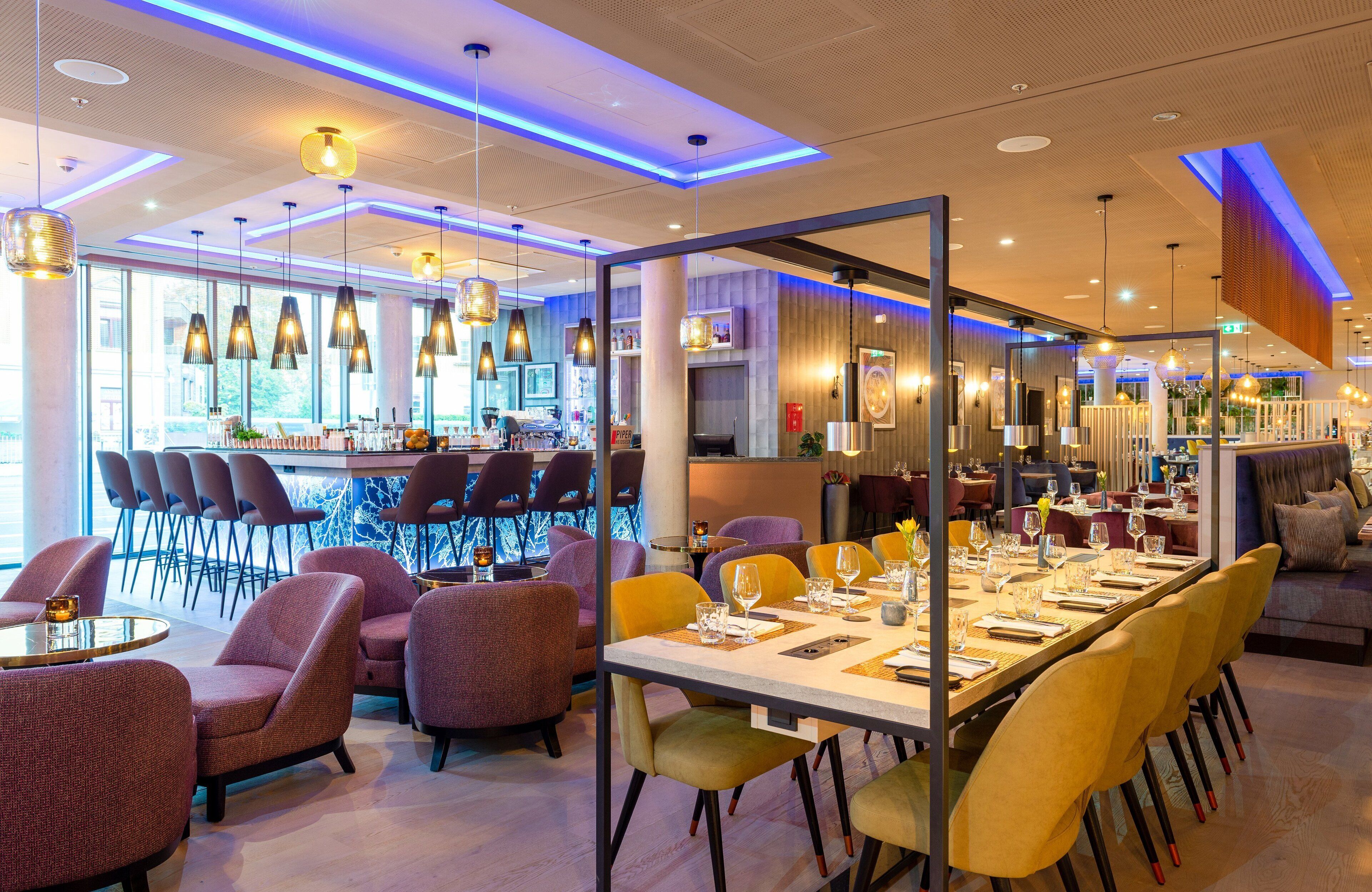 A modern restaurant with a long dining table, multiple chairs, and hanging pendant lights.