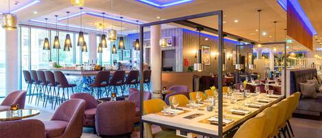 A modern restaurant with a long dining table, multiple chairs, and hanging pendant lights.