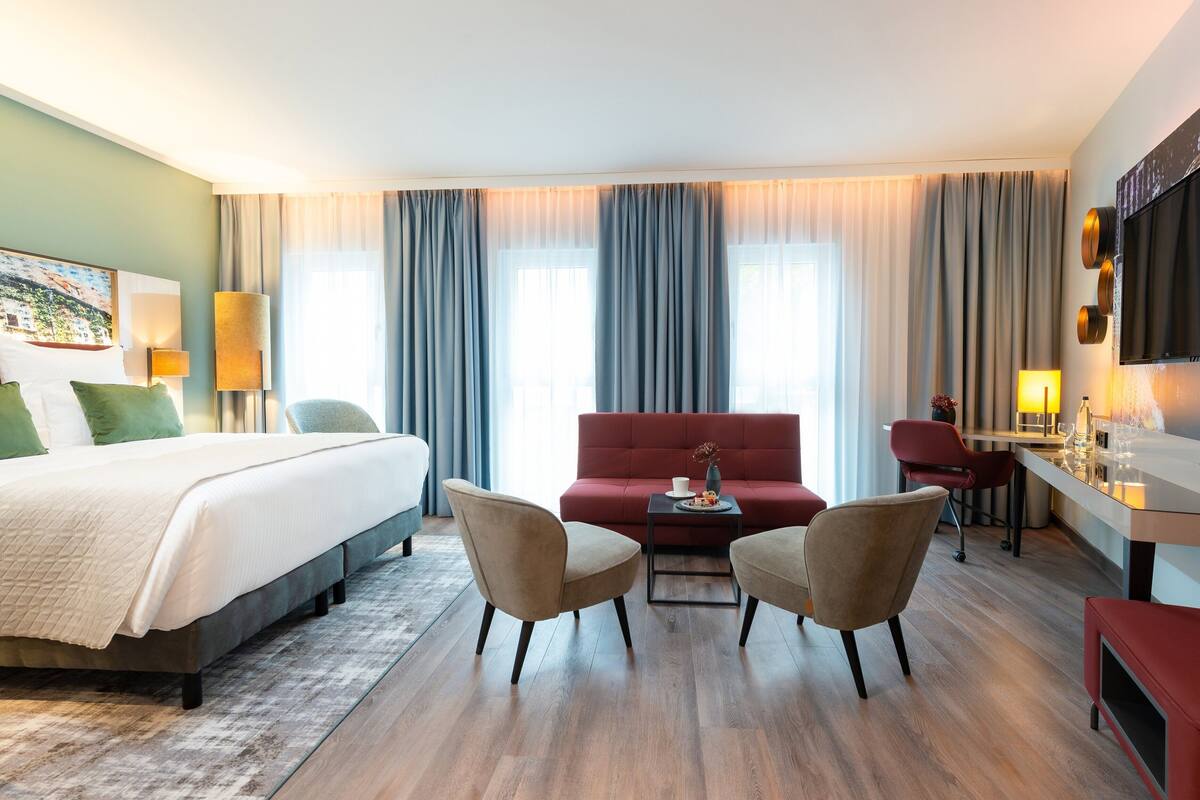 A hotel room with a bed, two armchairs, a red sofa, a small table with a vase, and a TV mounted on the wall.