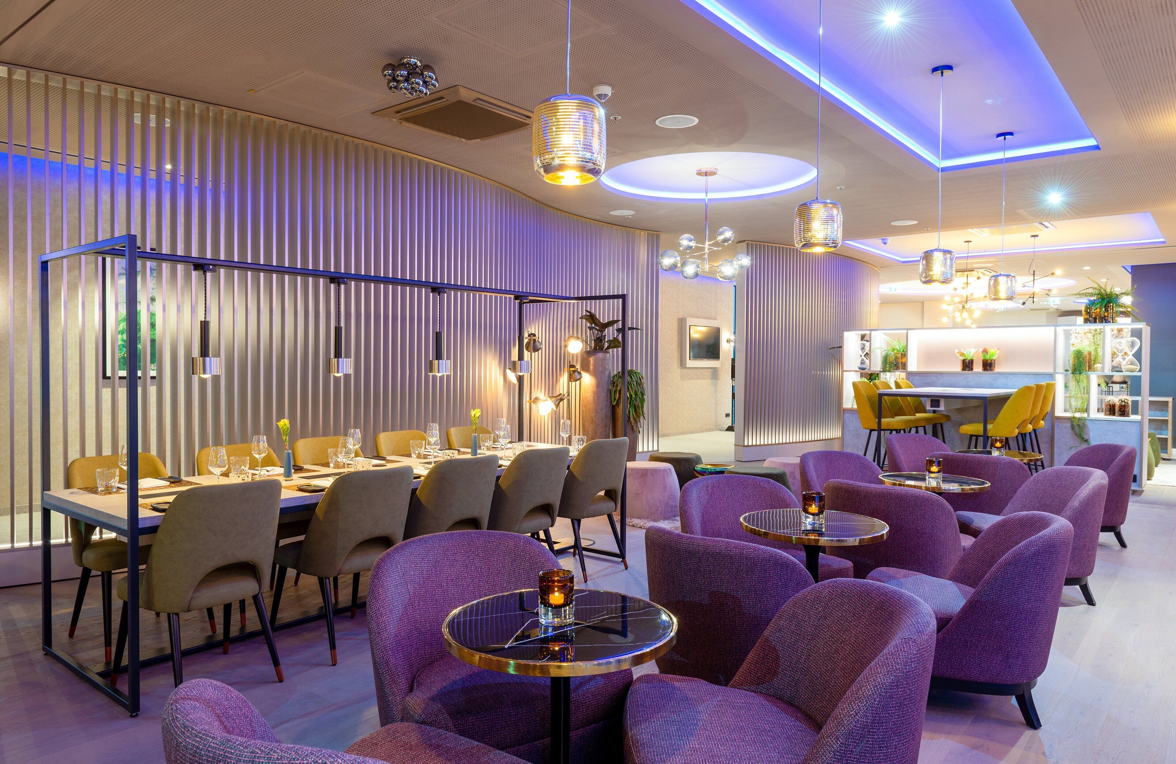 A modern dining area with a long table and purple chairs, a bar area with yellow chairs, and a kitchen in the background.