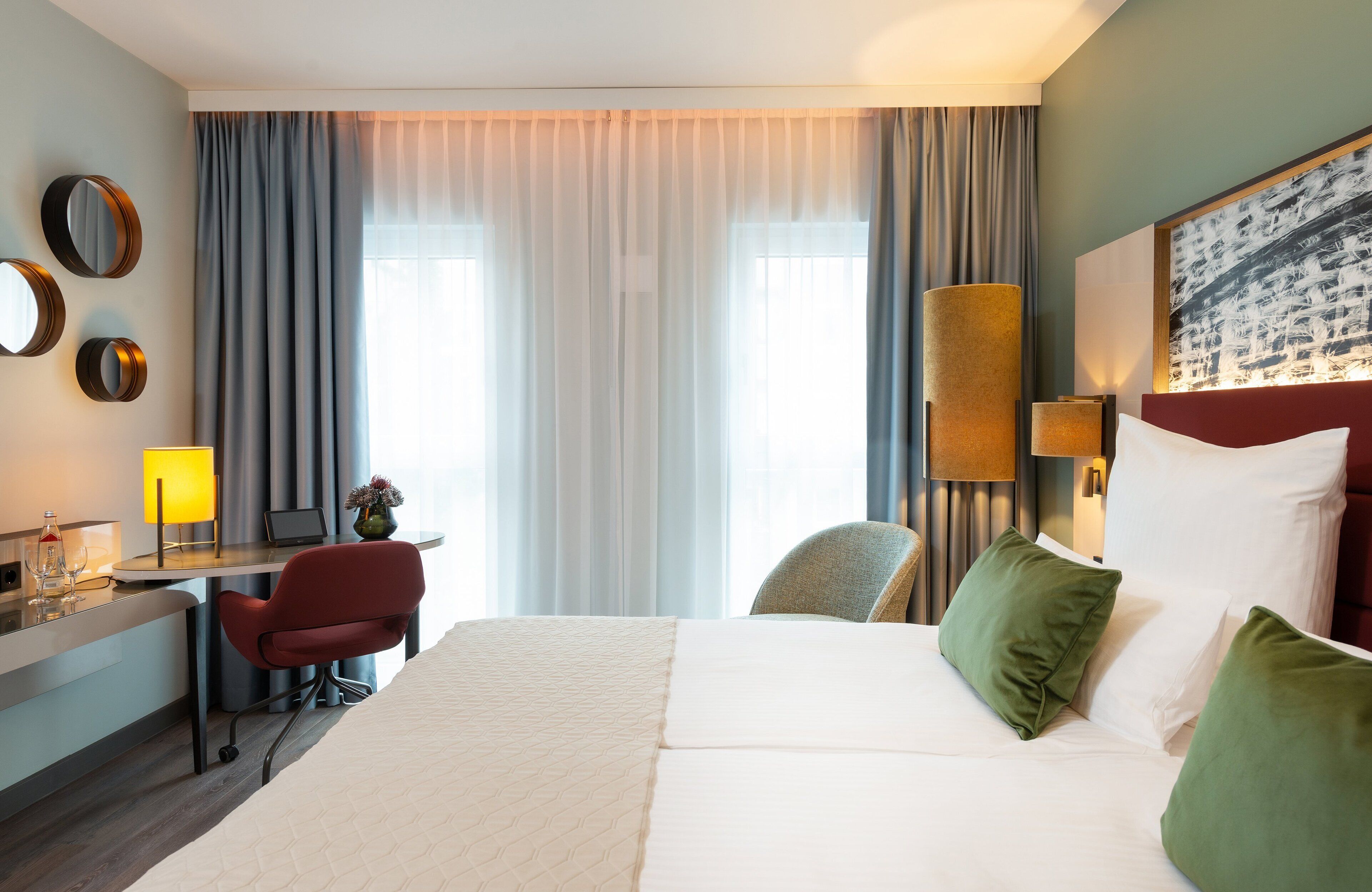 Habitación de hotel con una cama, un escritorio con lámpara, una silla y un cuadro grande en la pared.