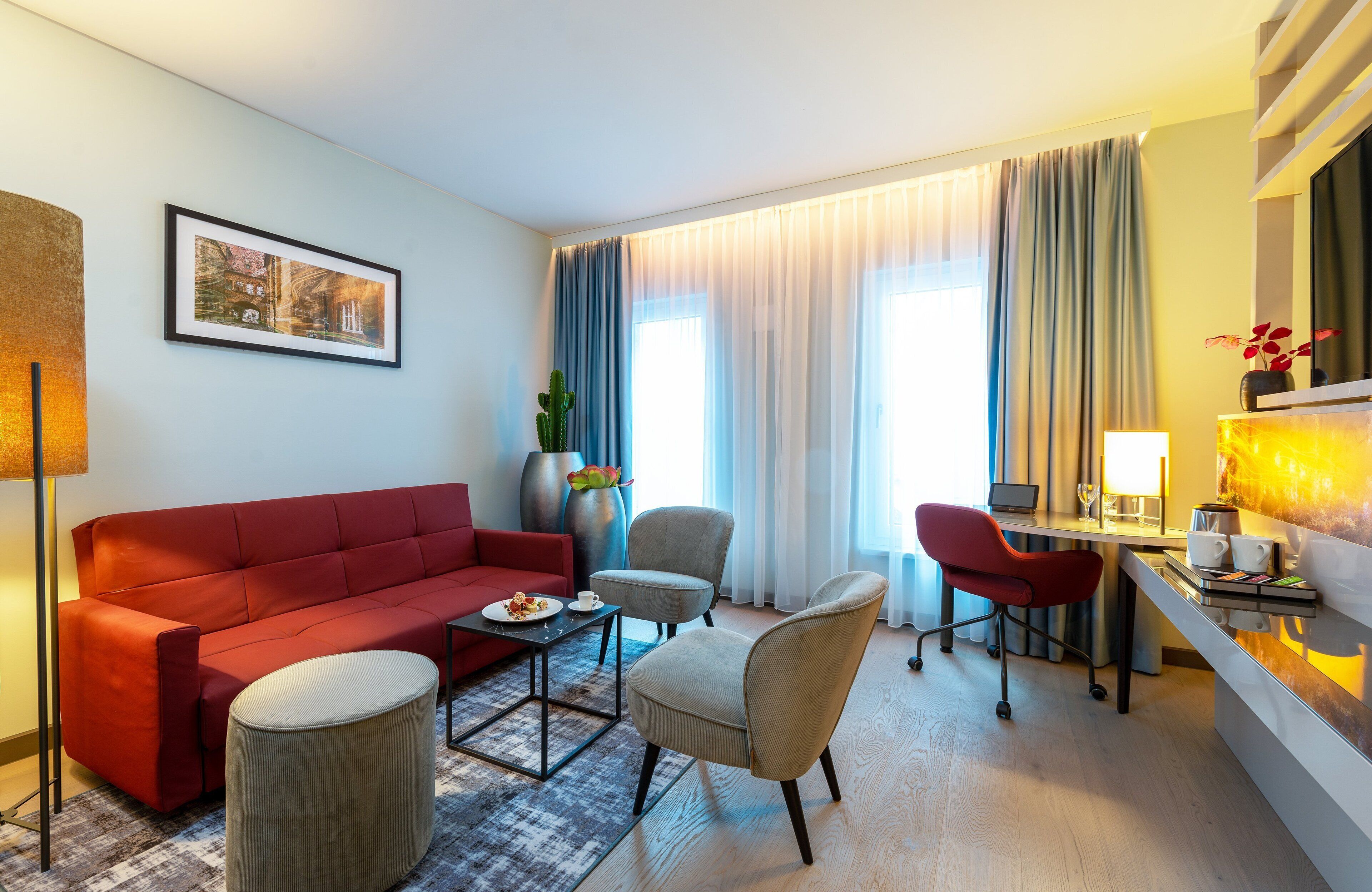 A modern hotel room with a red sofa, a grey armchair, a small round ottoman, a desk with a laptop, and a vase with flowers.