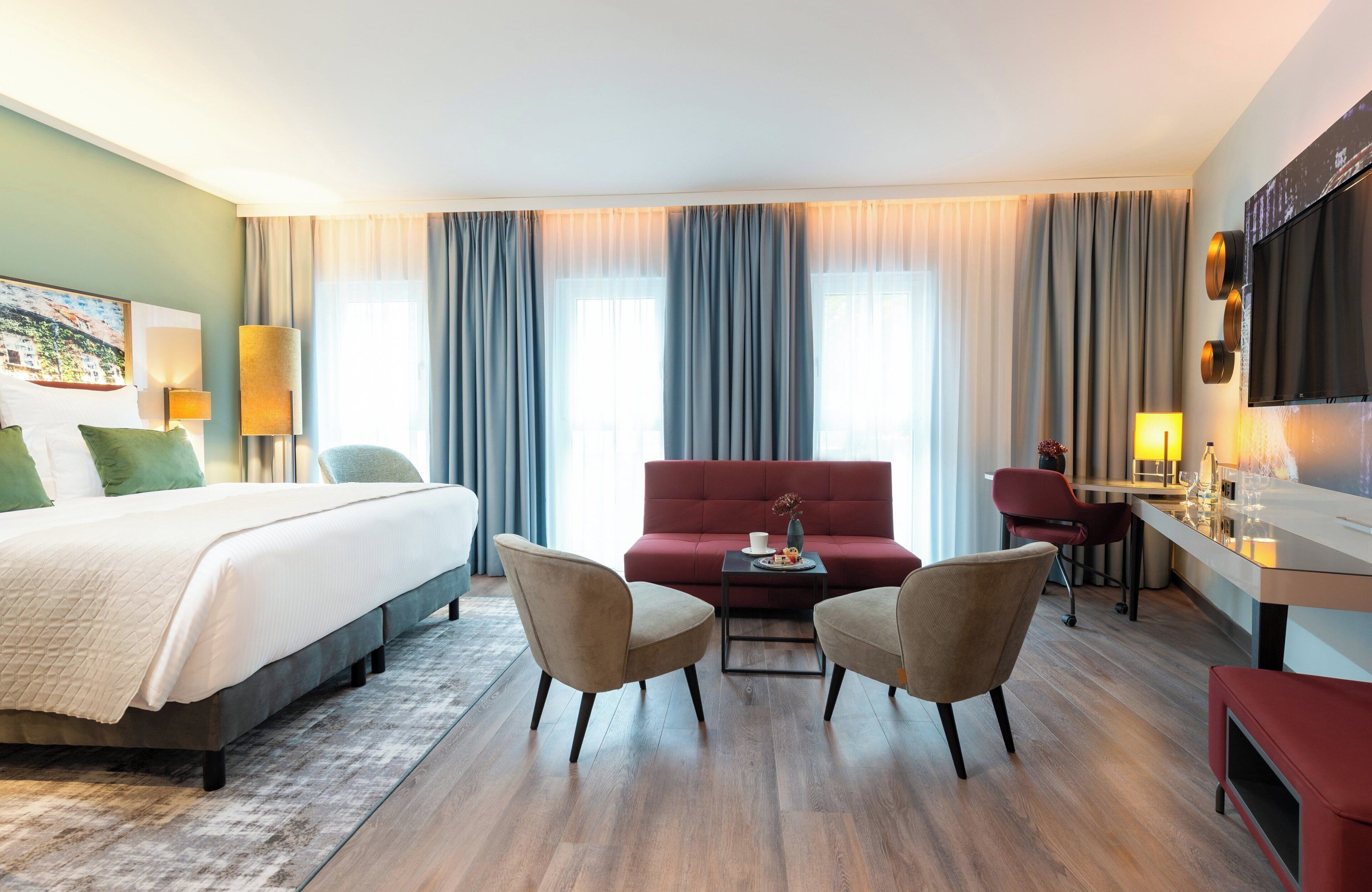 Ein Hotelzimmer mit einem Bett, zwei Sesseln, einem roten Sofa, einem kleinen Tisch mit einer Vase und einer Tasse sowie einem an der Wand befestigten Fernseher.
