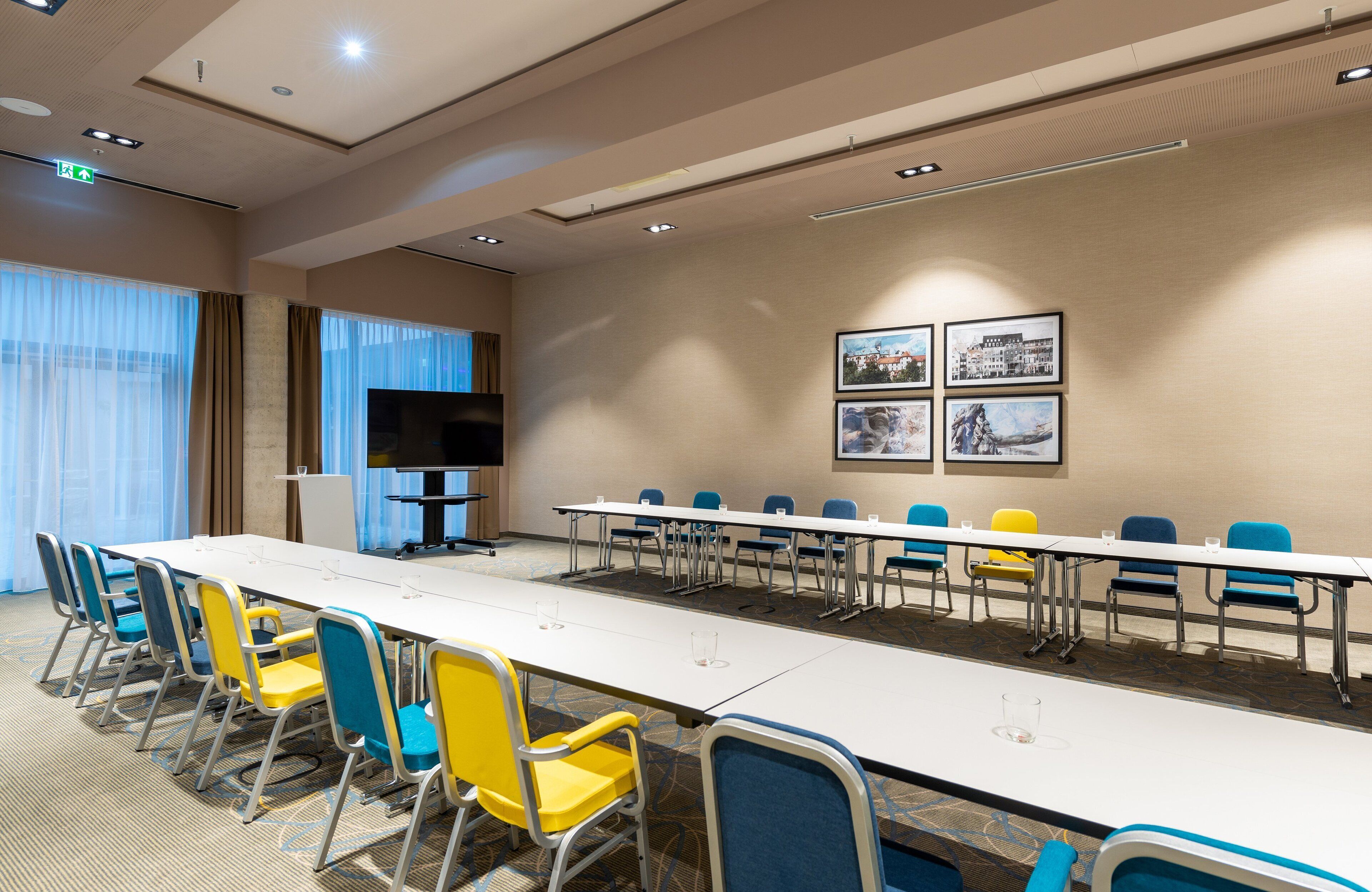 A conference room with a long table, blue and yellow chairs, a flat-screen TV, and framed pictures on the wall.