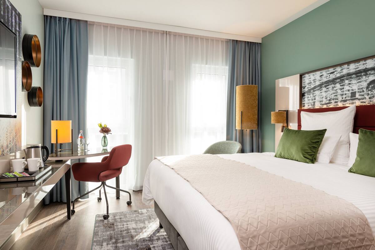 A hotel room with a bed, desk, chair, and a large window with curtains.