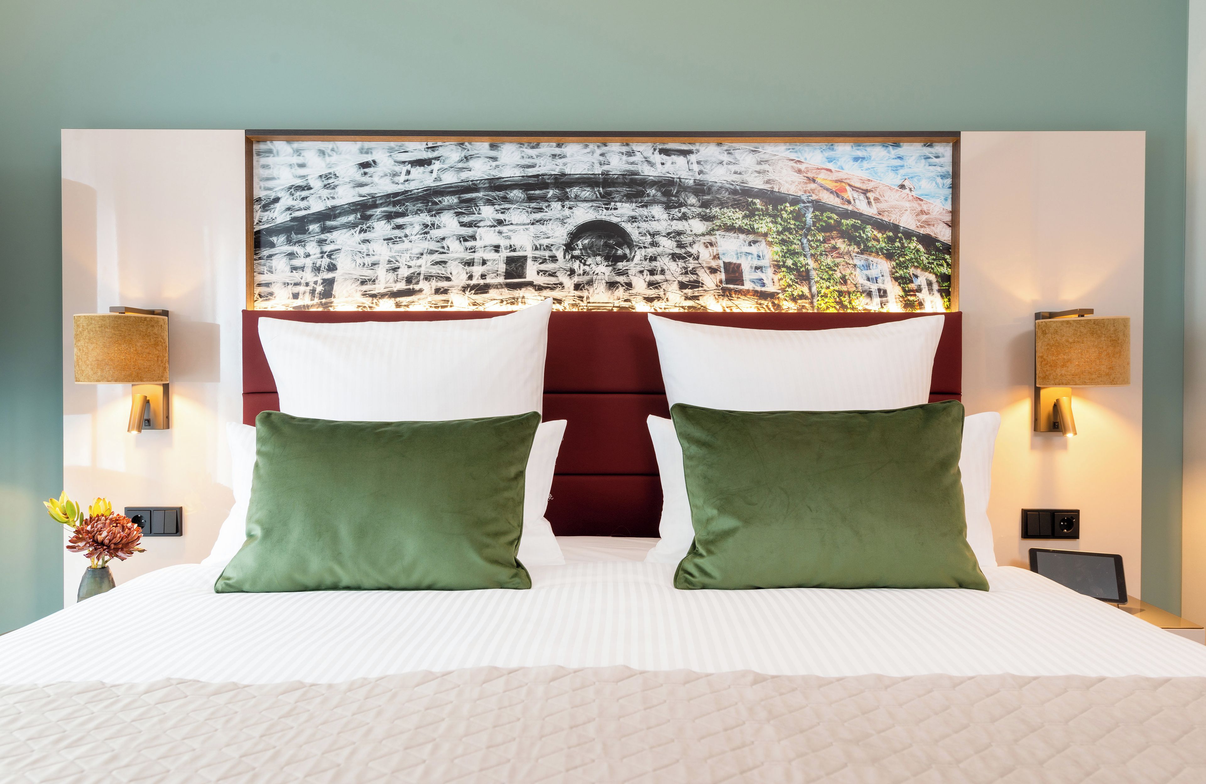 A hotel room with a large bed, two green pillows, a red headboard, and a cityscape view.