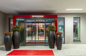 La entrada de un hotel con un marco rojo, plantas en macetas grandes y una alfombra roja con el nombre del hotel.