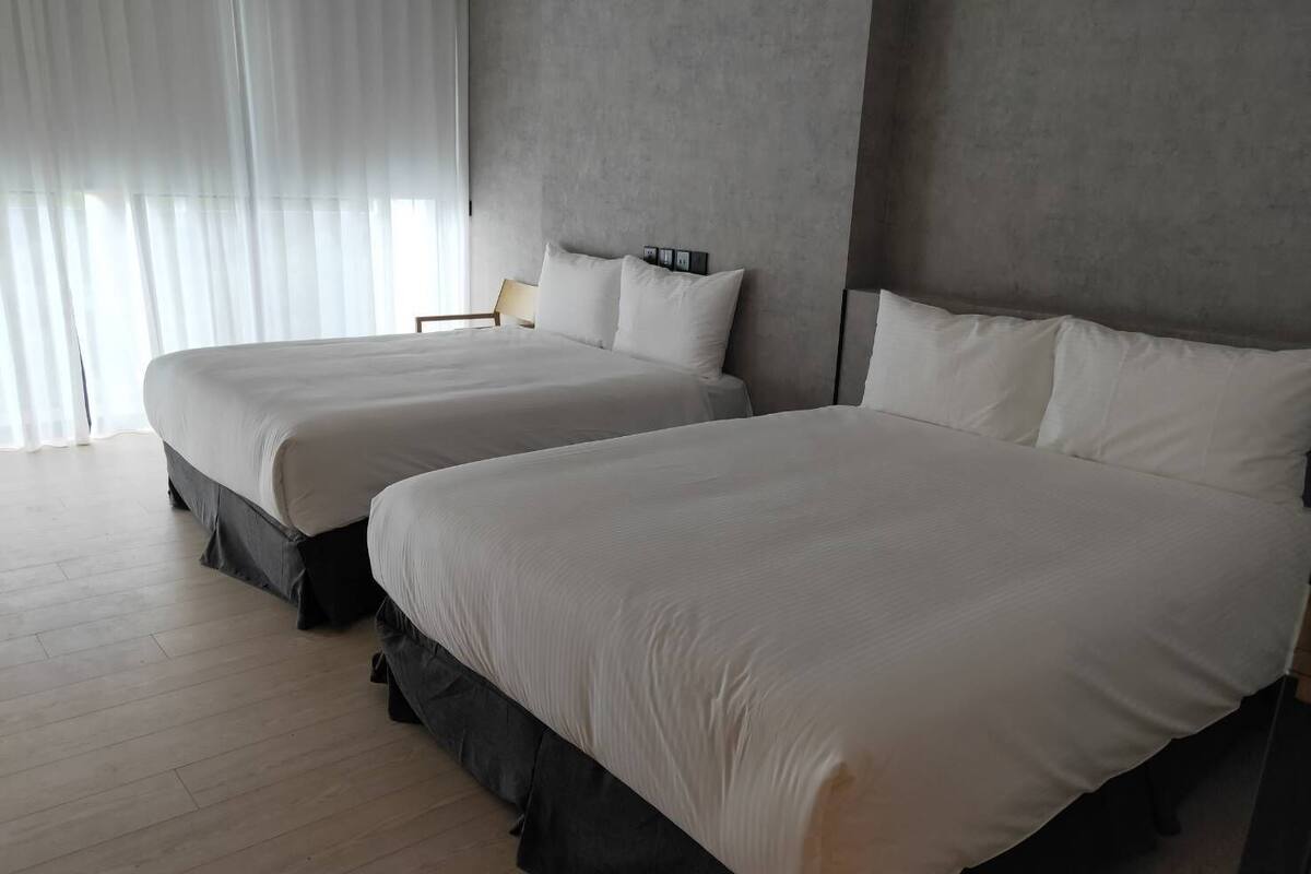 Una habitación de hotel con dos camas grandes, piso de madera y una ventana con cortinas translúcidas.