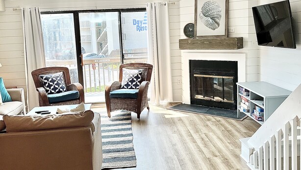 A spacious living room with a fireplace, a large window with a view of the outside, and a flat-screen TV mounted on the wall. - Bay views and Sunsets - 4 bedroom in the Heart of Dewey! (Dewey Beach)