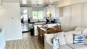 A modern kitchen with white cabinets, a dining area with a wooden table and chairs, and a living room with a sofa and a blue coffee table. - Bay views and Sunsets - 4 bedroom in the Heart of Dewey! (Dewey Beach)