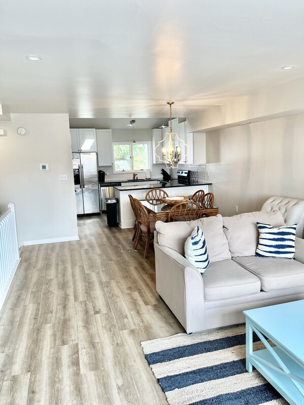 A modern kitchen with white cabinets, a dining area with a wooden table and chairs, and a living room with a sofa and a blue coffee table.