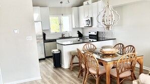 A modern kitchen with white cabinets, a black countertop, and a dining area with wicker chairs and a glass table. - Bay views and Sunsets - 4 bedroom in the Heart of Dewey! (Dewey Beach)