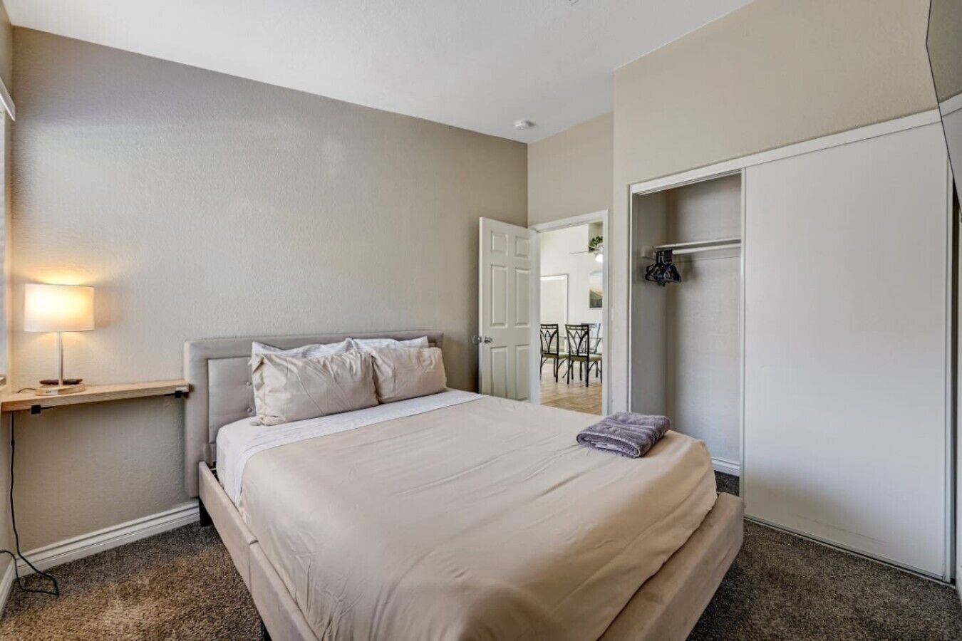 A hotel room with a bed, a bedside lamp, a wardrobe, and a door leading to a dining area.