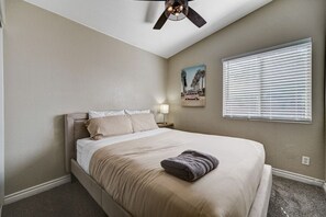 Un dormitorio con cama, ventilador de techo, ventana con persianas y un cuadro en la pared.
