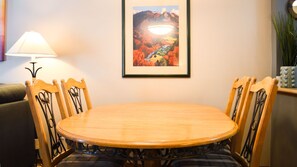 Een eethoek met een houten tafel en stoelen, een lijstwerk met een schilderij erboven, een lamp aan de linkerkant en een plant aan de rechterkant.