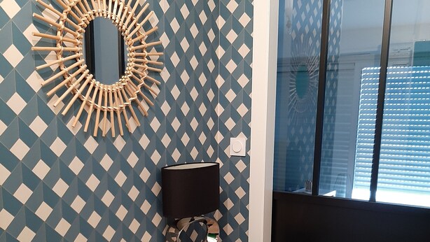 Ein Zimmer mit einer Wand mit geometrischem Muster, einem Holztisch mit Lampe, einem Spiegel und einem Fenster mit Jalousien.