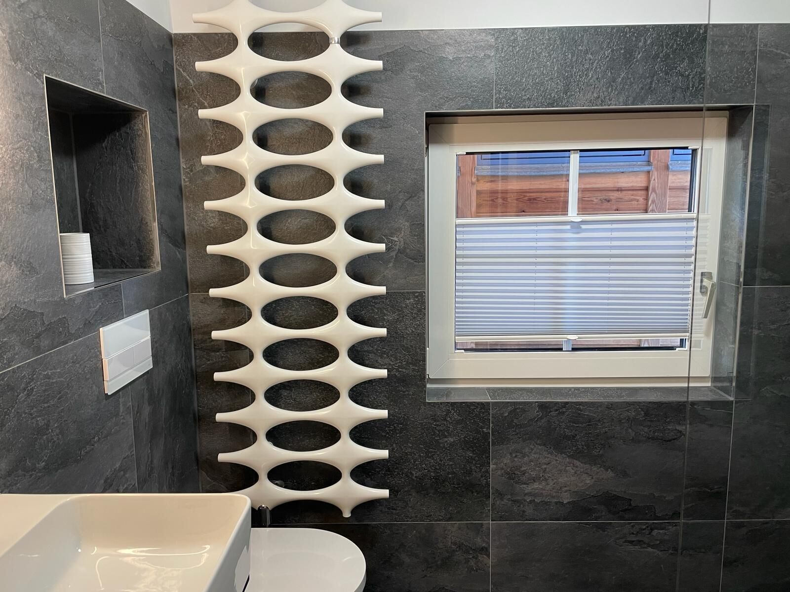 A modern bathroom with dark tiles, a white sink, a toilet, and a window with blinds.