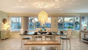 Uma sala de jantar com lustre, janelas amplas com vista para palmeiras e uma mesa posta para seis pessoas.
