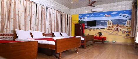 Quarto de hotel com duas camas de madeira, uma TV de tela plana, um ventilador de teto e um mural na parede retratando uma paisagem costeira.