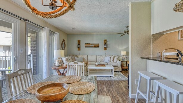 A spacious living room with a sofa set, a coffee table, and a dining area with a bench and table. There is a kitchen area with a countertop and bar stools. - Second row, three bed, two bath condo, complex pool (Surfside Beach)
