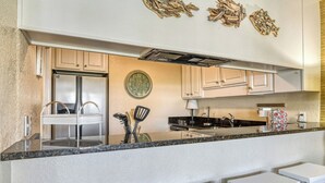 A modern kitchen with a stainless steel refrigerator, a clock, and white cabinets. There are three white bar stools in front of the counter. - Second row, three bed, two bath condo, complex pool (Surfside Beach)
