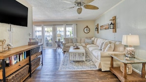 A spacious living room with a large sofa, a coffee table, and a TV mounted on the wall. There is a ceiling fan, a bookshelf, and a window with a view of the outside. - Second row, three bed, two bath condo, complex pool (Surfside Beach)