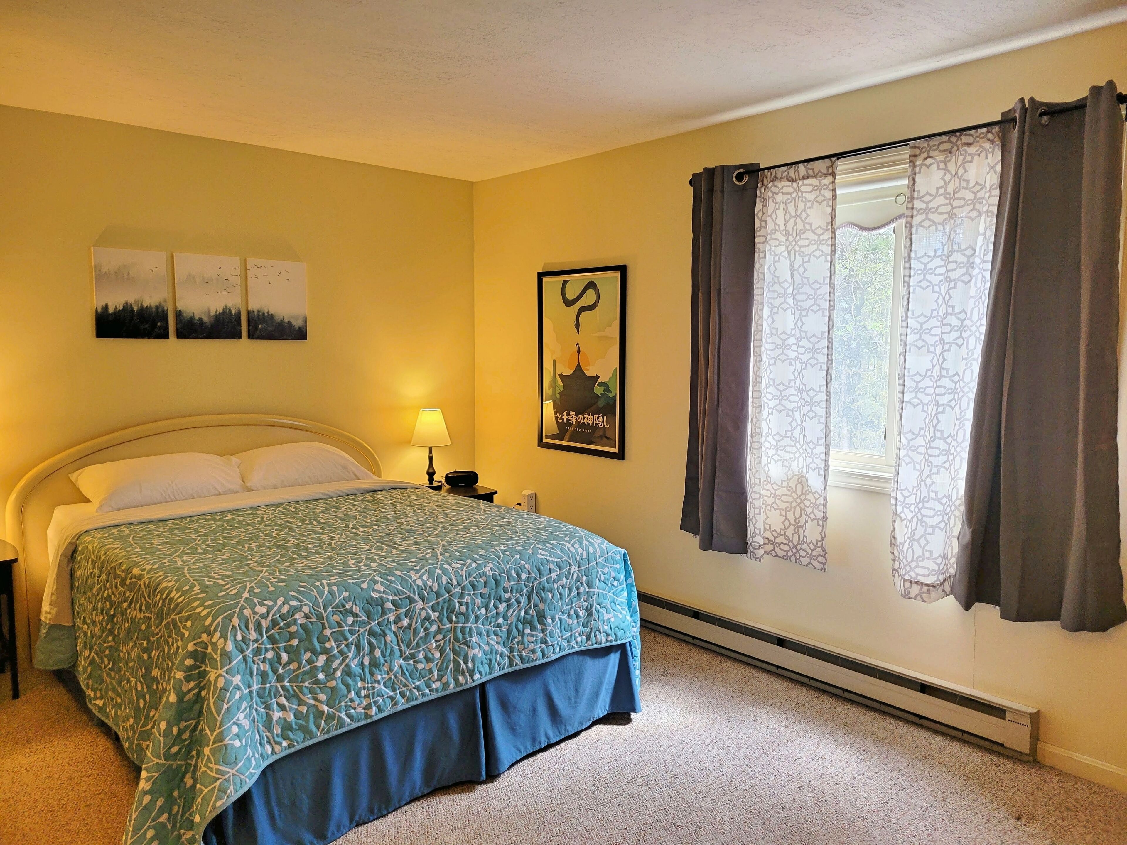 A bedroom with a bed, a nightstand, a lamp, a window with curtains, and wall art.