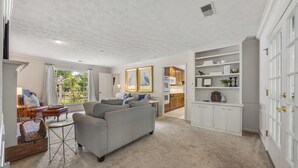 A spacious living room with a grey sofa, a small side table, and a wicker basket. There is a kitchen area with wooden cabinets and a white countertop visible through an open door. - Big Beds for Everyone!  Braves Stadium only 2.5 miles away! (Marietta)