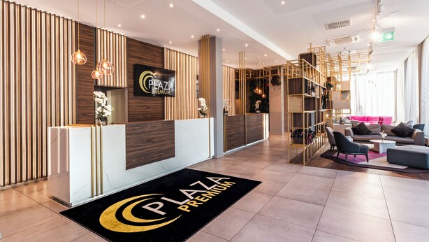 A modern reception area with a reception desk, a black and gold logo on the floor mat, and a seating area with sofas and a coffee table. - PLAZA Premium München (Munich)