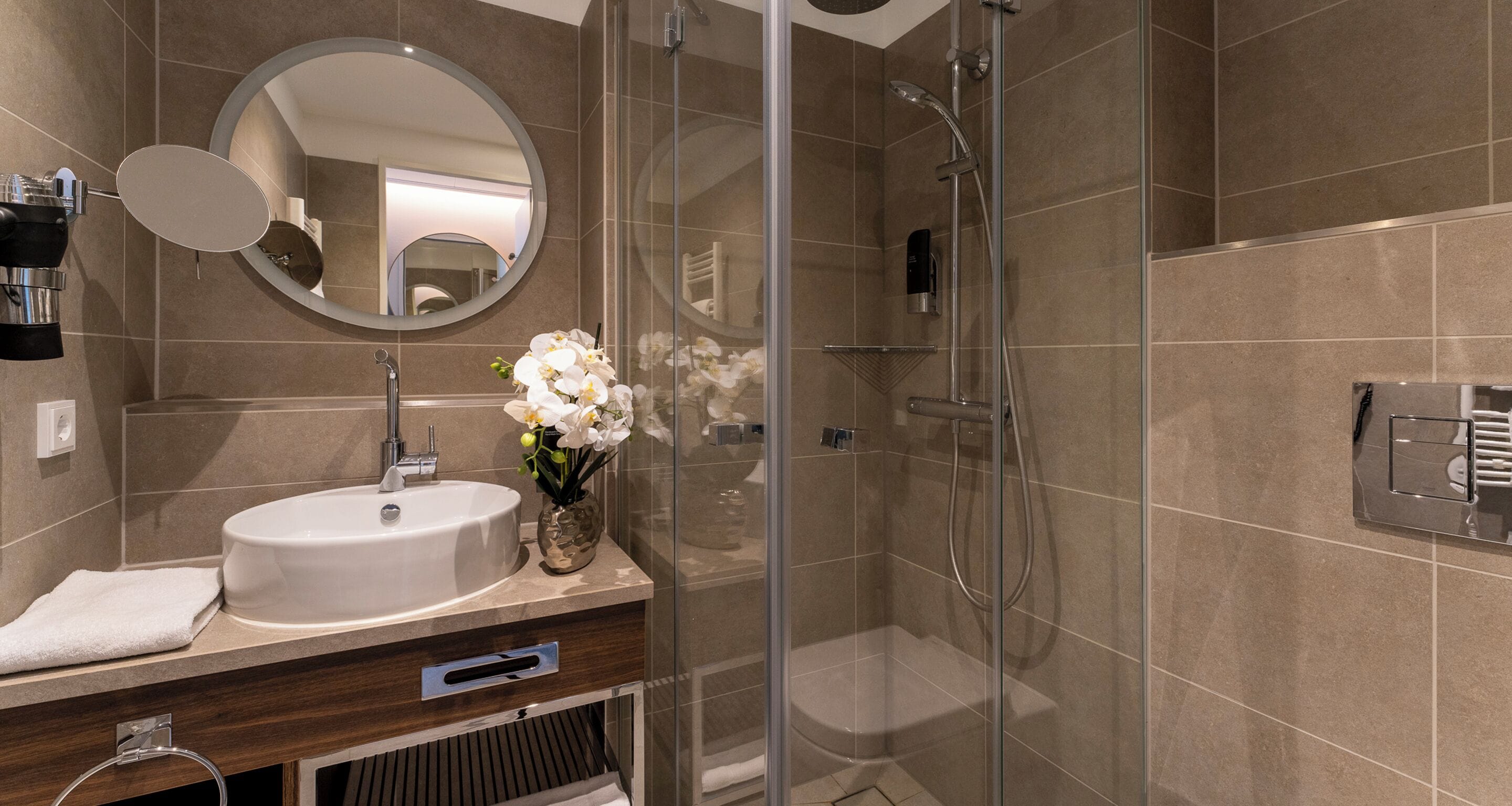 Un baño moderno con ducha de vidrio, un espejo redondo y un lavamanos blanco con un florero.