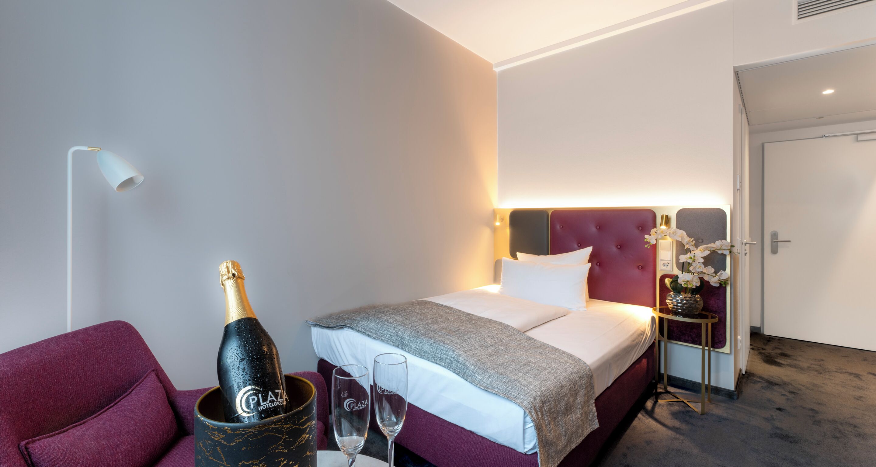 Une chambre d’hôtel avec un lit, une table de chevet ornée d’un vase de fleurs, une bouteille de champagne et deux verres.