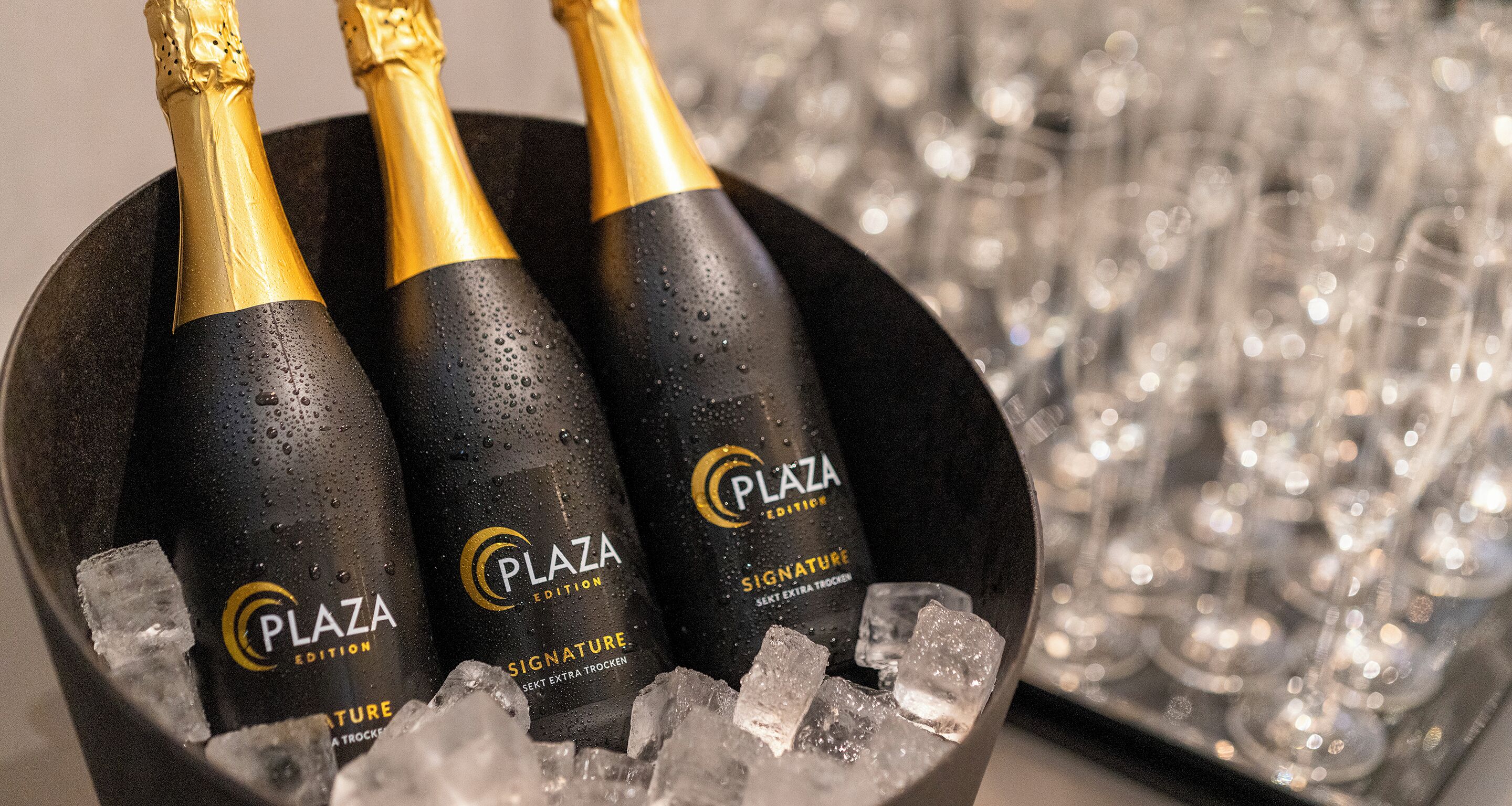 Three bottles of Plaza Signature champagne in a black bucket with ice cubes, surrounded by empty champagne glasses.