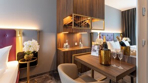 A hotel room with a bed, a small table with a bottle of champagne and glasses, a chair, and a mirror reflecting the room. - PLAZA Premium München (Munich)