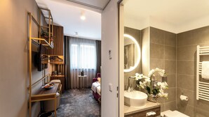 A hotel room with a wooden bookshelf, a small round table with a bowl of fruit, a bed, a bathroom with a sink, mirror, and a towel rack.
