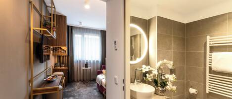 Una habitación de hotel con una estantería de madera, una mesita redonda con un frutero, una cama, un baño con lavabo, espejo y toallero.