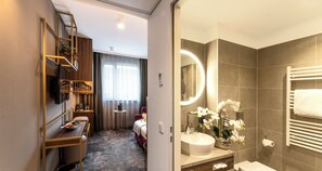 A hotel room with a wooden bookshelf, a small round table with a bowl of fruit, a bed, a bathroom with a sink, mirror, and a towel rack.