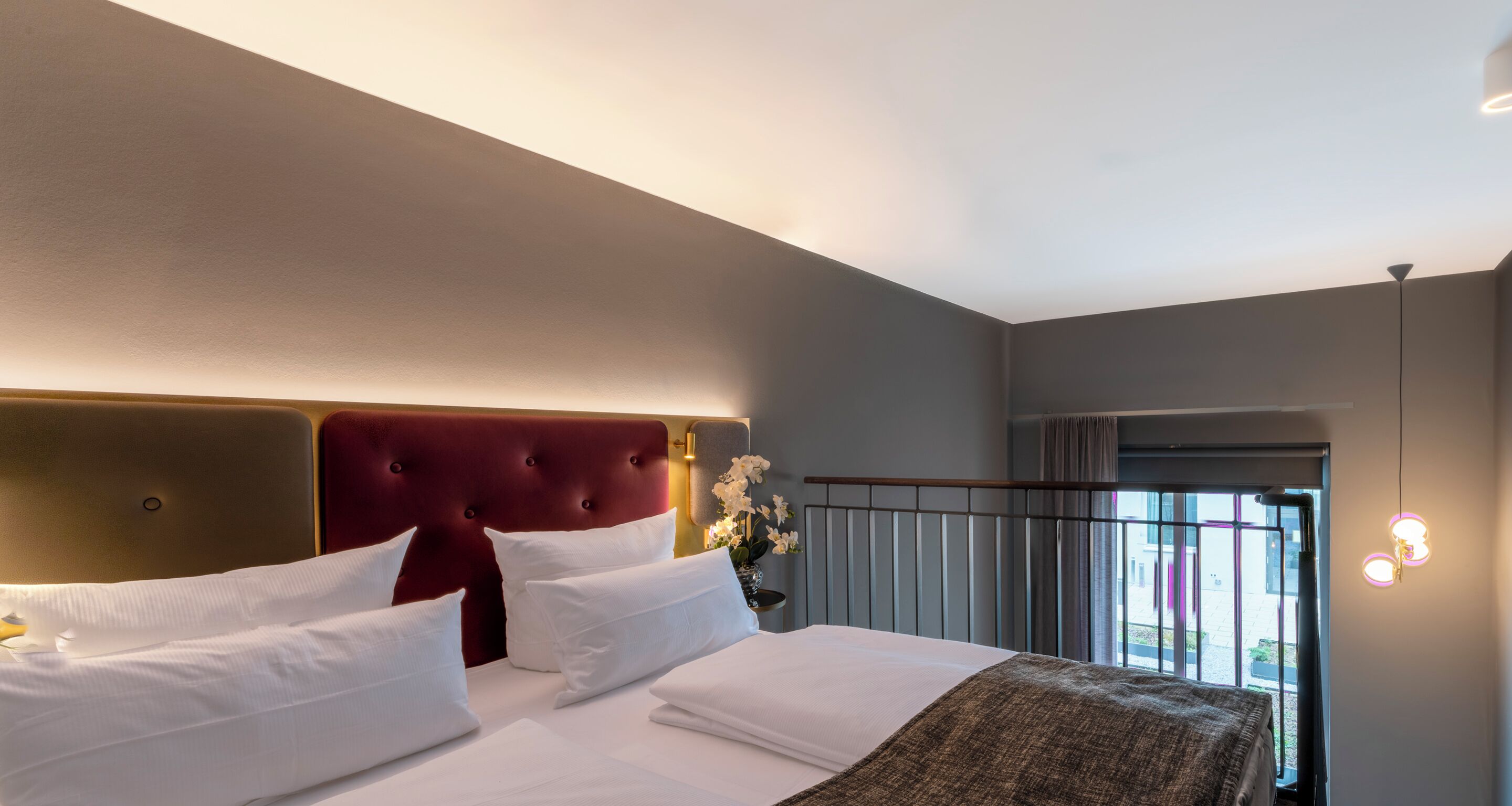 A hotel room with a large bed, a red upholstered headboard, white bedding, and a view of the outside through a window.