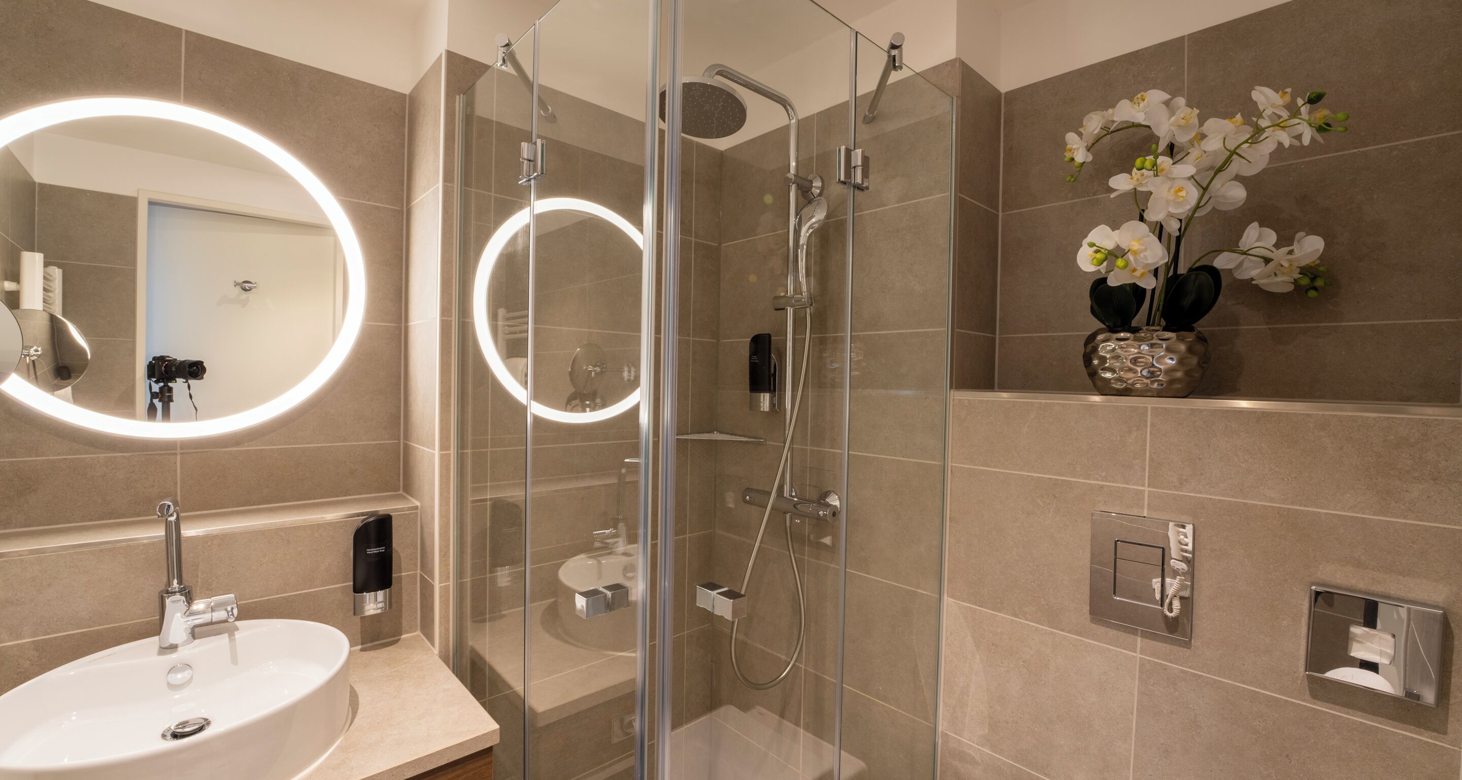 Un baño moderno con ducha envuelta en vidrio, un espejo redondo con iluminación, un lavabo y un jarrón con orquídeas blancas.