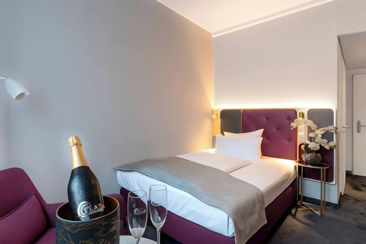 A hotel room with a bed, a bedside table with a vase of flowers, a bottle of champagne, and two champagne flutes.