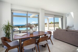 A modern living room with a dining area, large windows, and a view of the sea.