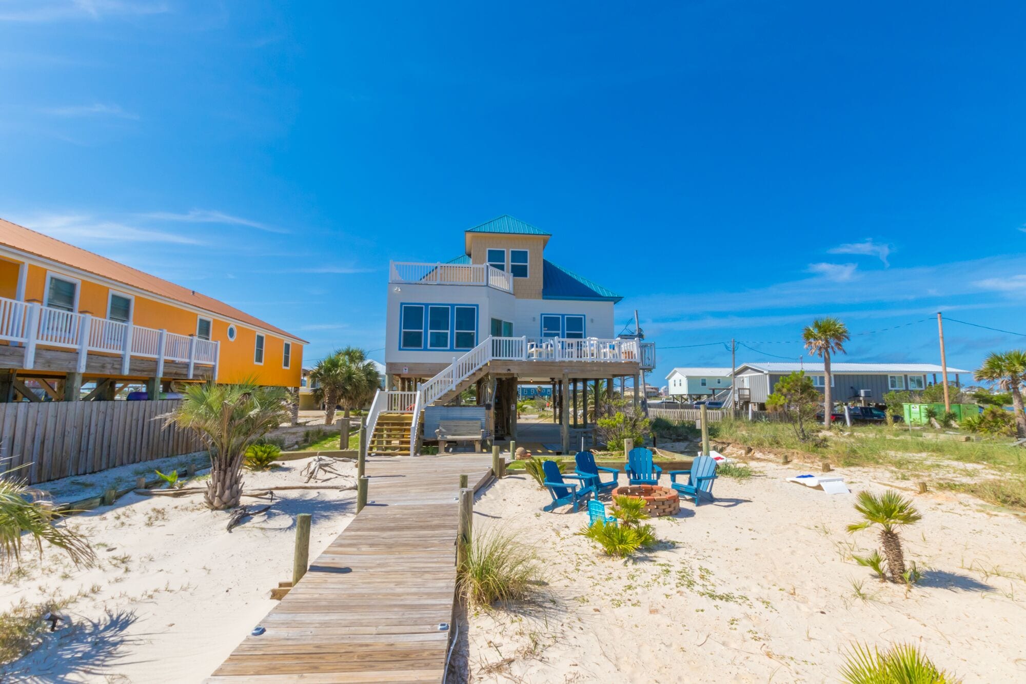 Ein direkt am Strand gelegenes Anwesen mit einer Holzterrasse, einer blauen Sitzgruppe im Freien und einem zweistöckigen Gebäude mit Balkon.