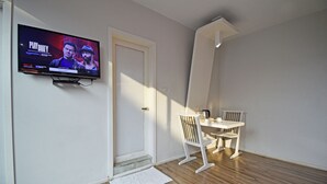Quarto com televisão na parede, porta branca, mesa pequena com cadeira e chão de madeira.