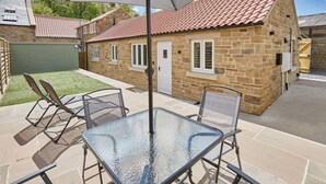 Une maison en pierre avec un toit de tuiles, une table en verre avec des chaises et une pelouse verdoyante.