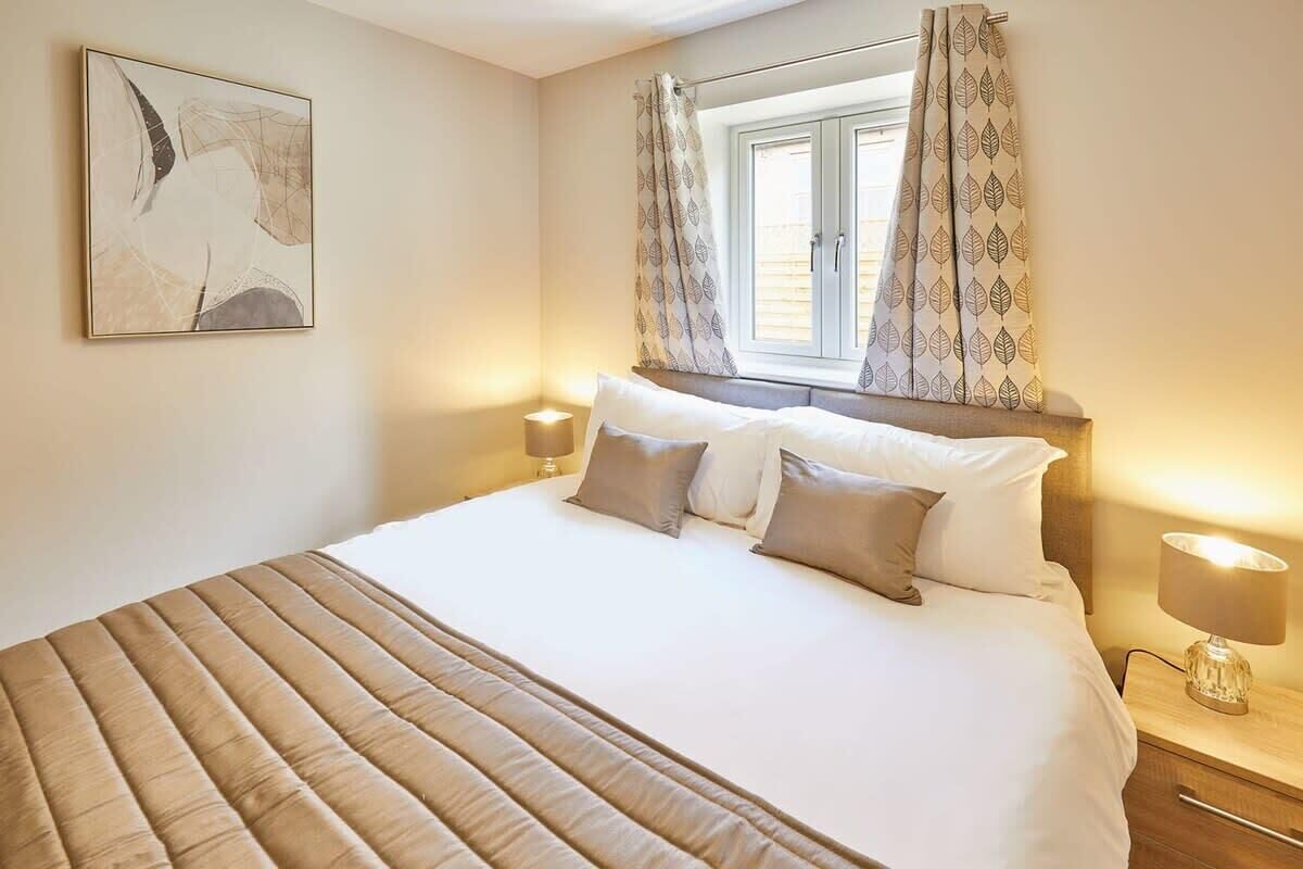 Un lit bien fait, recouvert d’une couverture à rayures, avec deux lampes de chevet et une fenêtre ornée de rideaux à motifs.