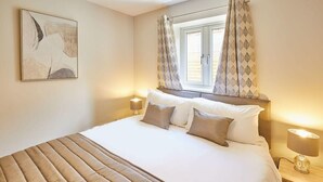 Un lit bien fait, recouvert d’une couverture à rayures, avec deux lampes de chevet et une fenêtre ornée de rideaux à motifs.