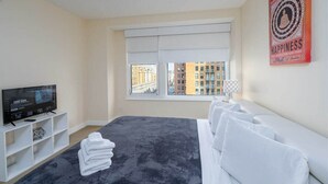 Une chambre d’hôtel avec un lit, une télévision, une fenêtre donnant sur des immeubles et un mur orné d’une affiche.
