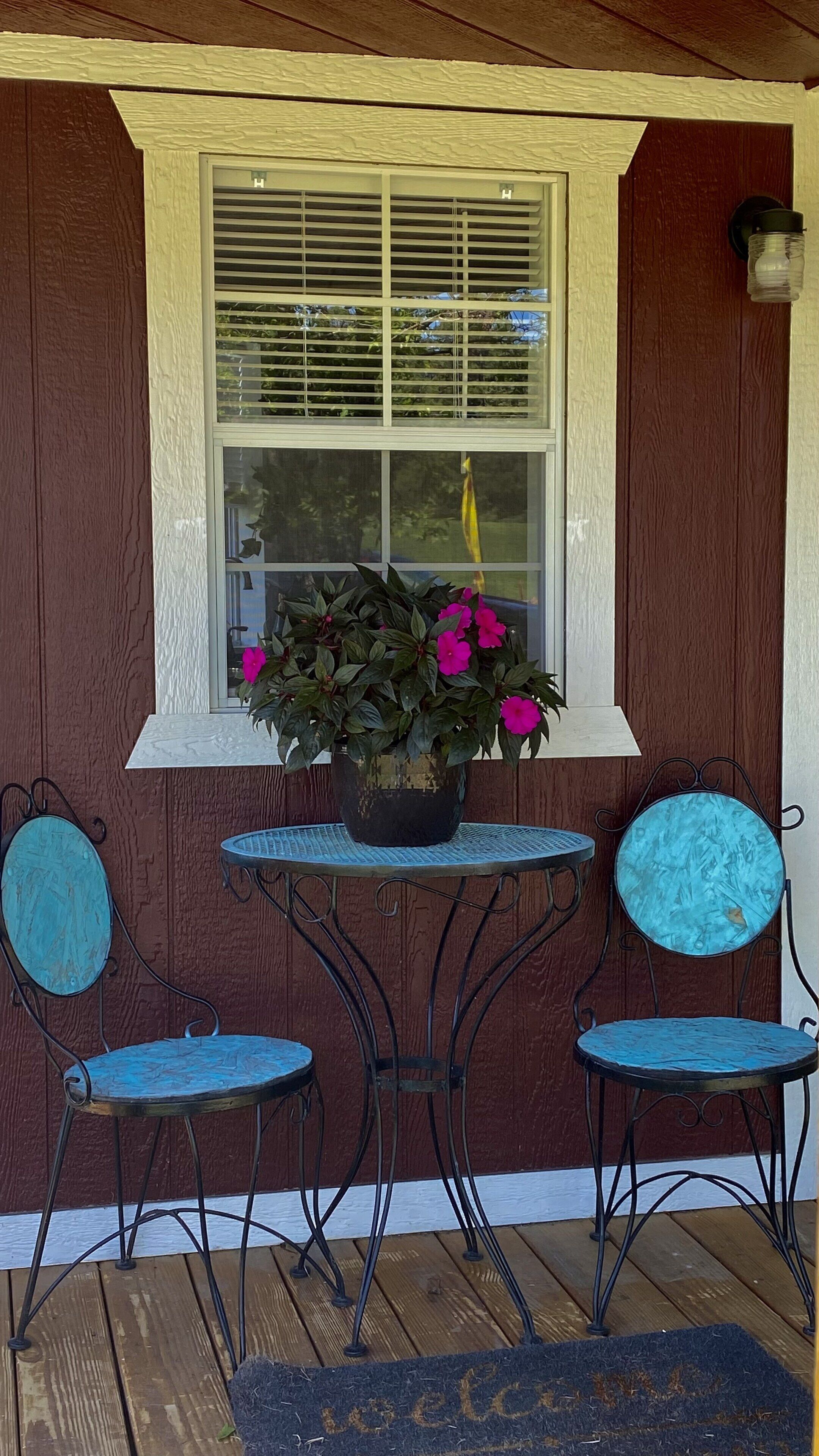 Un porche con una mesa de madera y dos sillas, una planta en maceta con flores rosadas y una ventana con persianas.