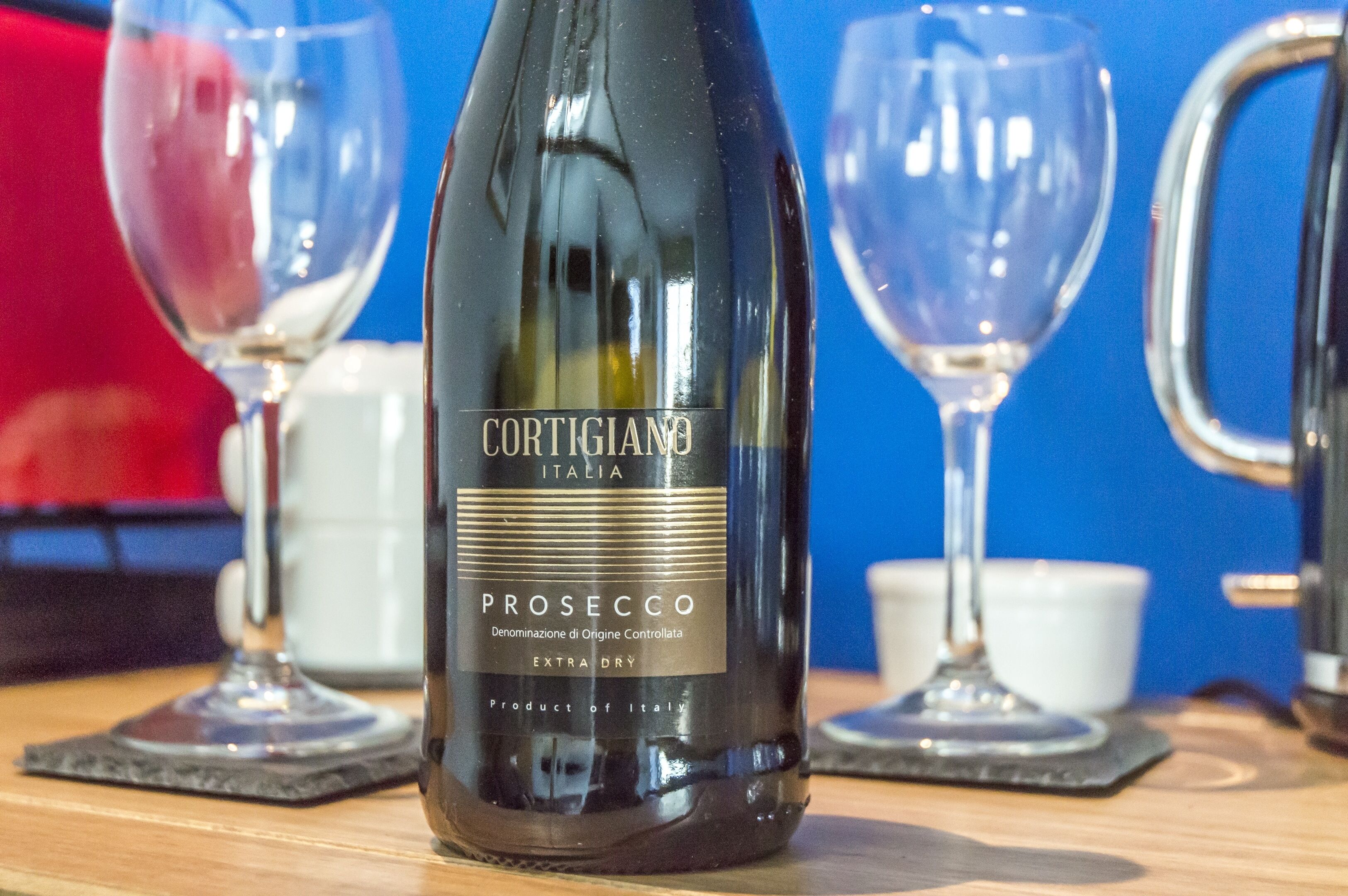 Una botella de Cortigiano Prosecco sobre una mesa de madera, con dos copas de vino y una taza blanca al fondo.